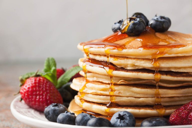 Close-up delicious pancakes, with fresh blueberries, strawberries and maple syrup on a light background. With copy space. Sweet maple syrup flows from a stack of pancake.