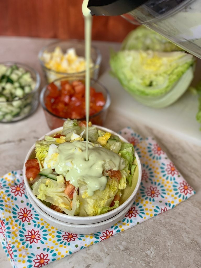 salad in white bowl sitting on top of a colorful napkin with salad fixings and lettuce in the back with avocado ranch dressing being poured on