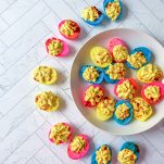 plate of multi colored deviled eggs that are blue, pink, and yellow.; deviled eggs surrounding half of the plate as well, filled with egg yolk mixture