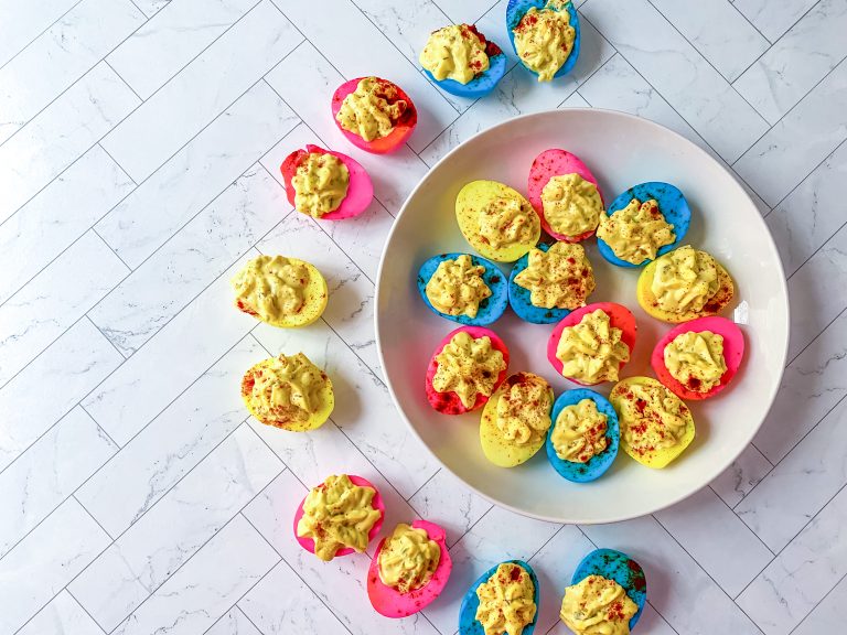 plate of multi colored deviled eggs that are blue, pink, and yellow.; deviled eggs surrounding half of the plate as well, filled with egg yolk mixture
